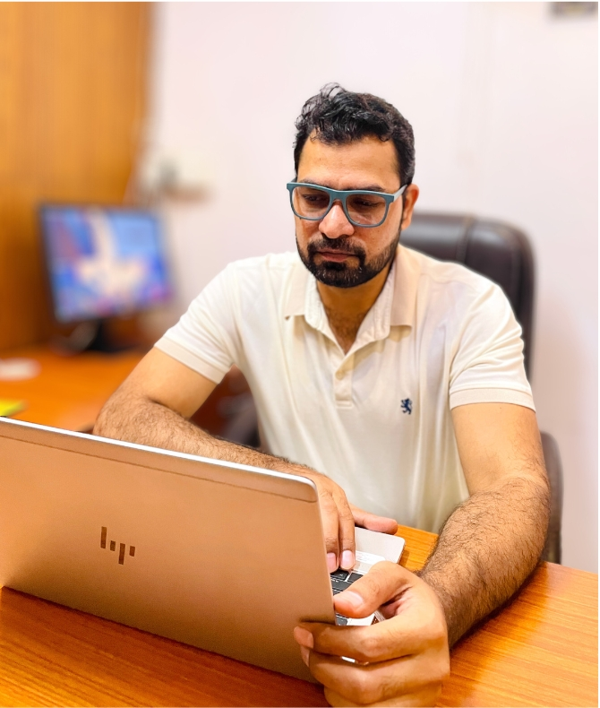 A person in a light polo shirt sits at a desk, typing on a silver HP laptop, with a blurred computer monitor in the background.